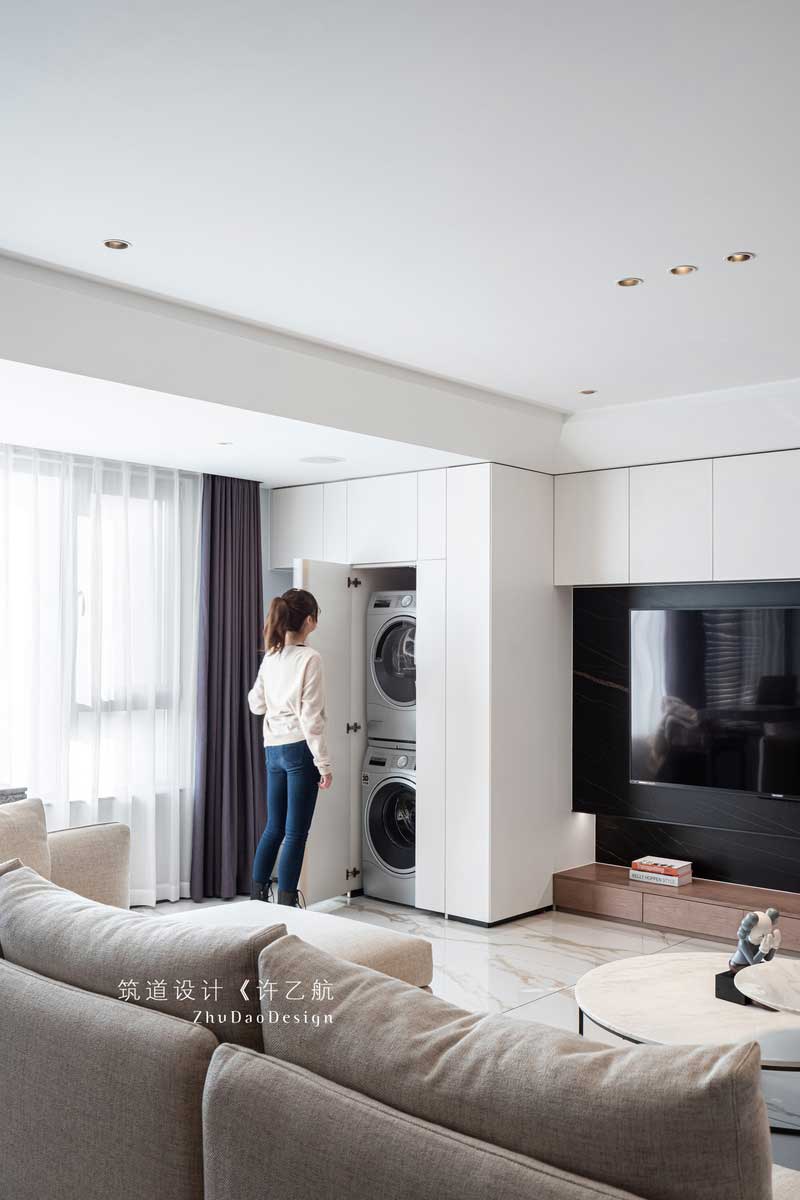 設(shè)計師充分利用陽臺的空間，把橫在客廳與陽臺的墻體打通，讓整個陽光通過大面積的窗戶照射進(jìn)來，使整個空間采光通透；又將電視背景墻與之結(jié)合，并做了隱形門處理，將洗衣機(jī)、烘干機(jī)嵌入，既美觀又實(shí)用。